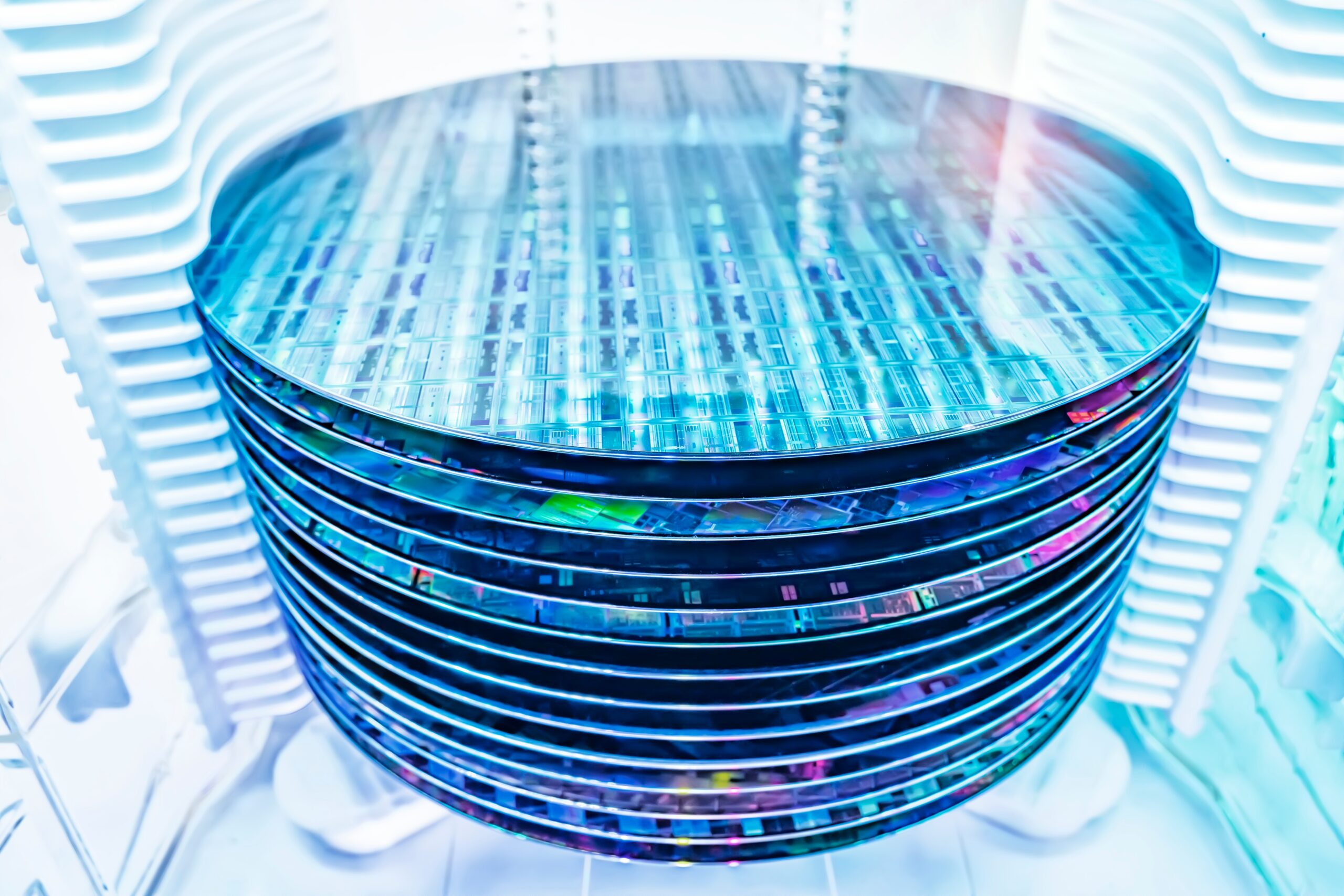 A stack of shiny silicon wafers, each reflecting colorful light, is shown in a bright, high-tech manufacturing environment, likely used for the production of semiconductors or microprocessors.