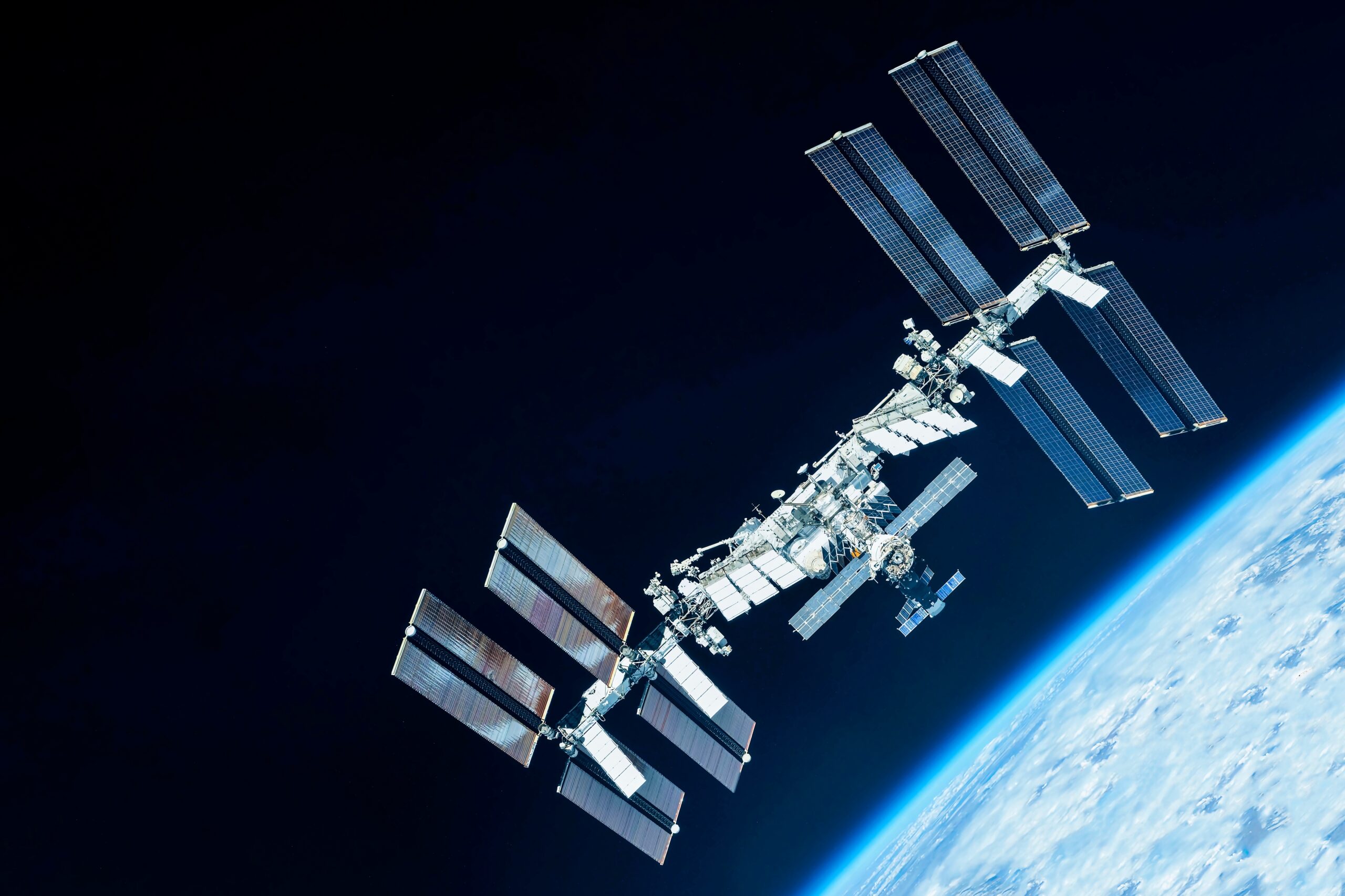 The International Space Station is orbiting above Earth, with its large solar panels deployed against the blackness of space and the blue curve of the planet visible below.