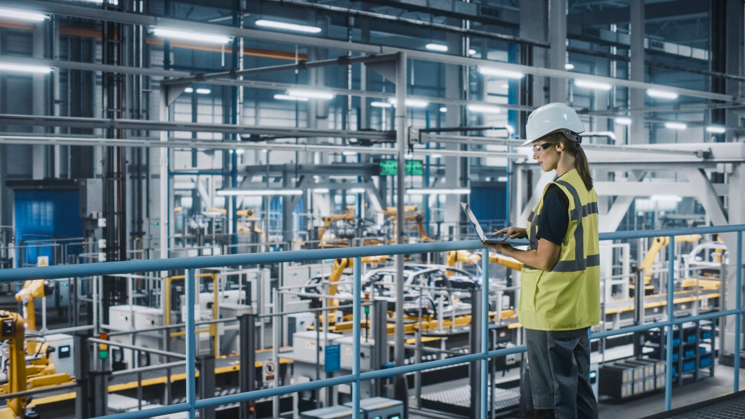 A person wearing a safety helmet and a high-visibility vest stands on a platform in a modern factory. They are using a laptop to monitor or control the robotic machines and automated assembly lines below.